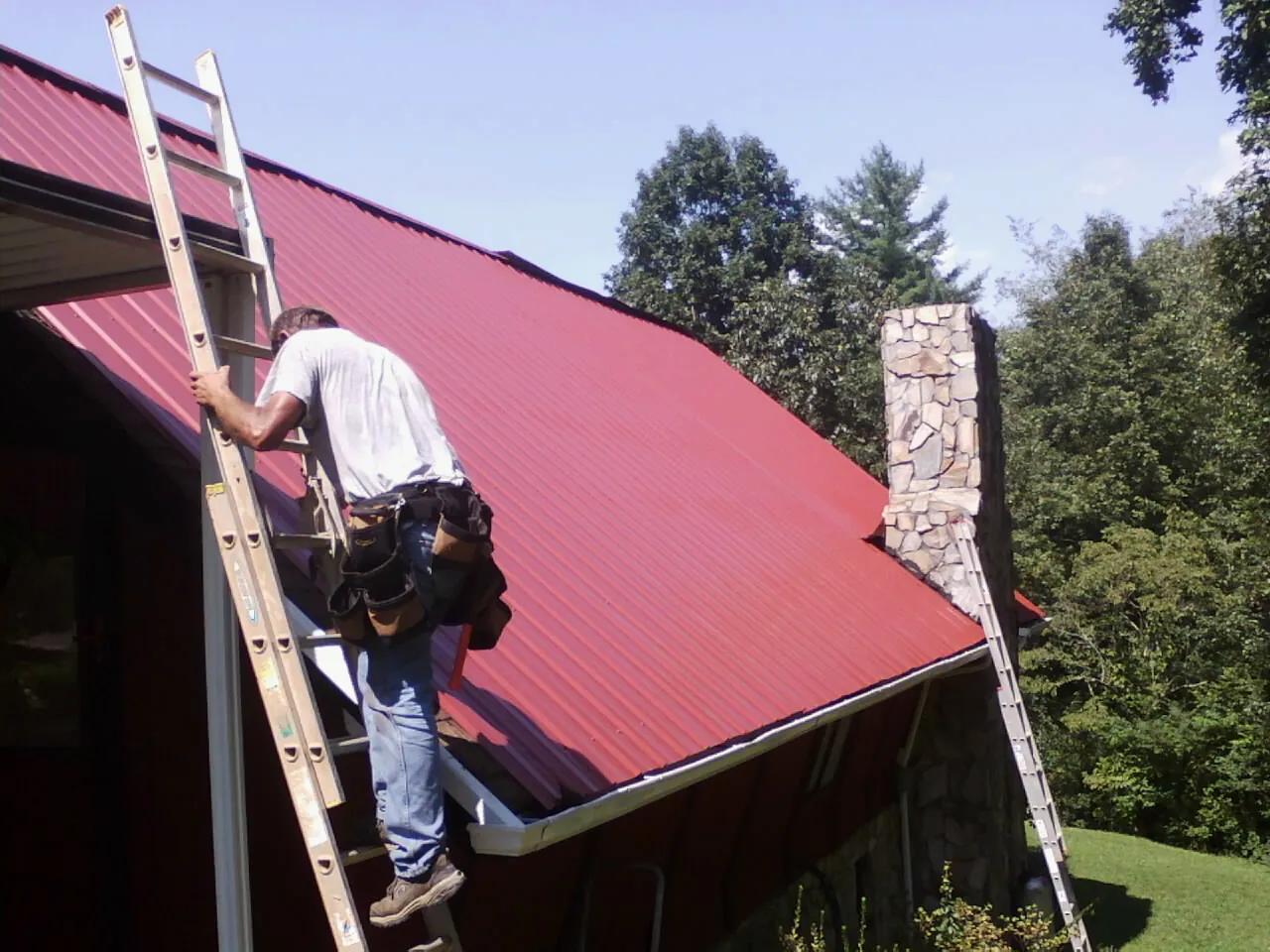 Roofer on a ladder beside a red metal roof and stone chimney — Soffit & Fascia Repair by Roofing Brothers in Morehead City
